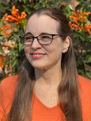 A middle-aged white woman with very long light brown hair looks slightly to one side while smiling. She is wearing glasses and an orange v-neck t-shirt. Out of focus in the background is a brick wall covered in flame vines: dark green leaves and orange flowers.